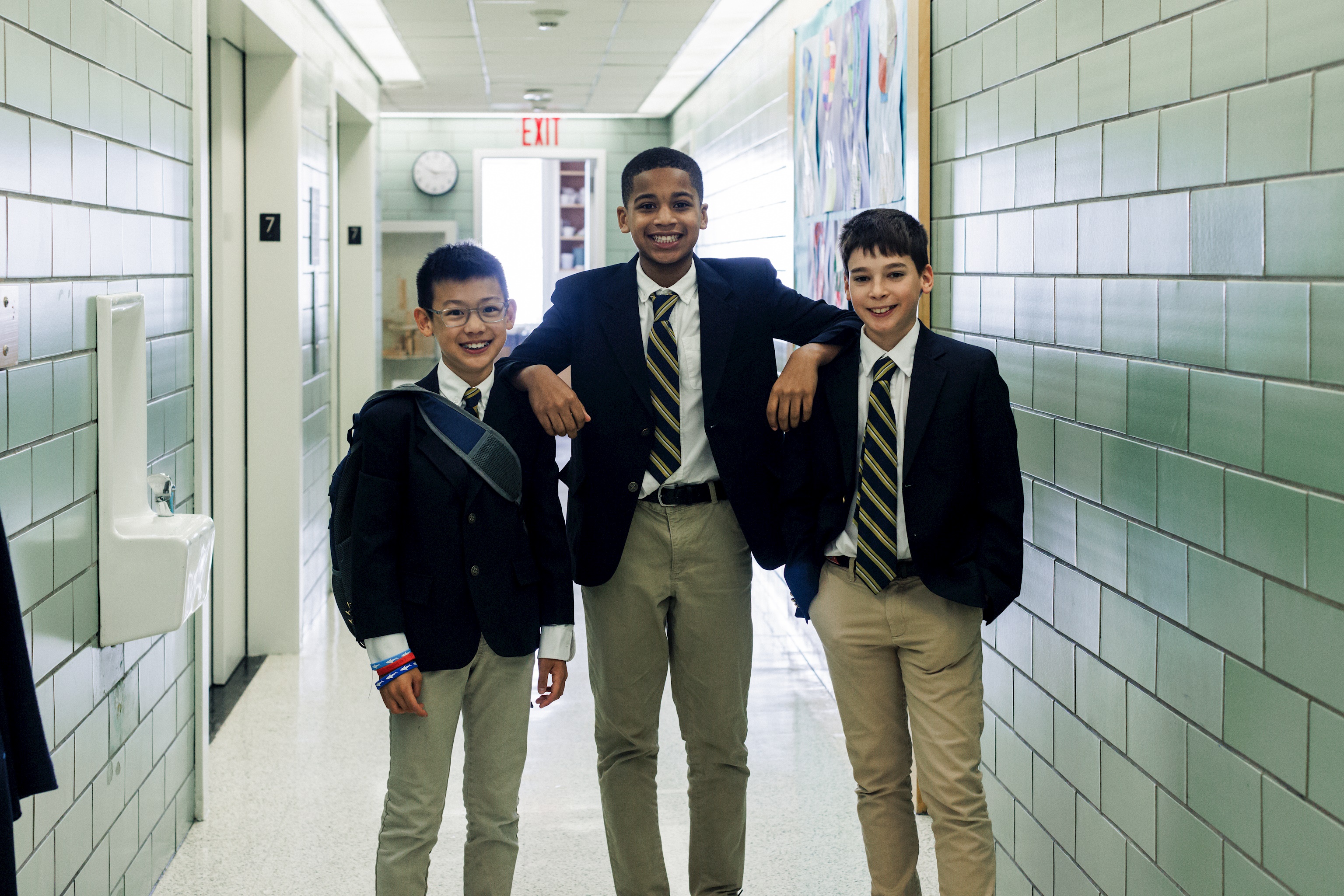 Students in school uniforms standing in a hallway, with middle school boys waiving and girls in the background.